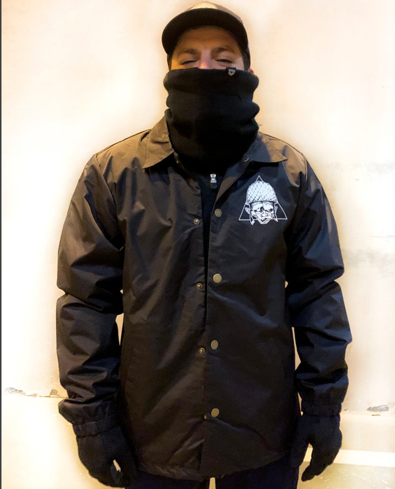 May include: A black nylon jacket with a snap closure and a white graphic of a skull wearing a helmet inside a triangle.