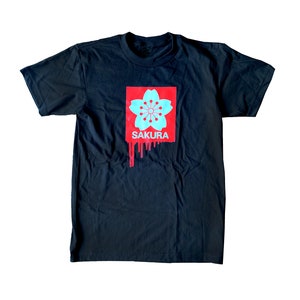 May include: Black t-shirt featuring a red and blue graphic of a cherry blossom flower with the word "SAKURA" printed below it.