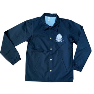 May include: A black coach jacket with a white skull and triangle design on the left chest. The jacket has a snap closure and two pockets.