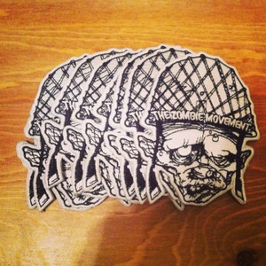 Könnte beinhalten: Ein Set mit sechs schwarzen und weißen gestickten Patches, die einen Zombie mit einem Militärhelm zeigen. Die Patches haben den Text "The Zombie Movement" auf dem Helm.