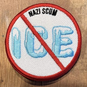 Op de afbeelding: Een ronde patch met een witte achtergrond, een rode rand en een rode diagonale lijn. De patch toont de zwarte woorden "NAZI SCUM" boven het blauwe woord "ICE" met een druipend ijs-effect.