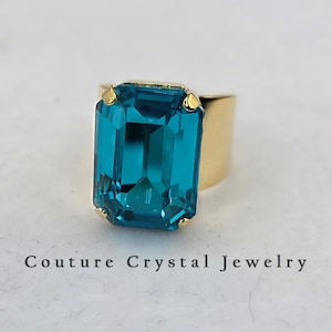 May include: A gold-toned ring featuring a large, rectangular, emerald-cut teal crystal held by four prongs. The band is wide and smooth. The text Couture Crystal Jewelry is printed in black below the ring on the light-colored surface.