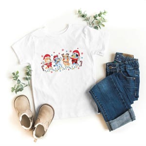 May include: White short-sleeved t-shirt featuring a festive graphic of cartoon characters dressed in Santa hats and holiday attire. The design includes candy canes and string lights. The shirt is paired with blue denim jeans and tan boots.