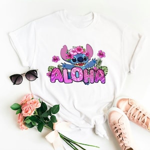 Puede incluir: Camiseta blanca con un personaje de dibujos animados azul que lleva una corona de flores rosas y la palabra "ALOHA" en letras rosas con un patr&oacute;n floral.