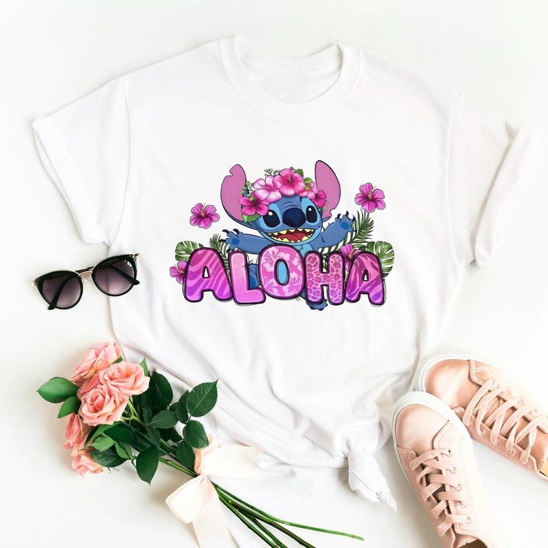 Puede incluir: Camiseta blanca con un personaje de dibujos animados azul que lleva una corona de flores rosas y la palabra "ALOHA" en letras rosas con un patr&oacute;n floral.