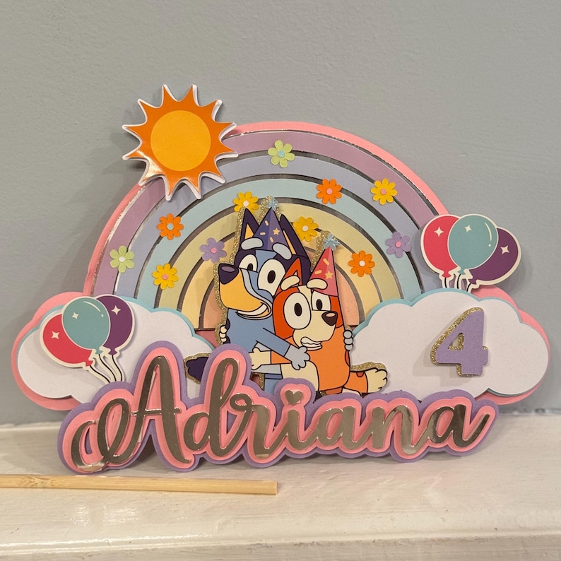Może przedstawiać: Kolorowy topper na tort z dwoma kresk&oacute;wkowymi psami, tęczą, słońcem i imieniem "Adriana" z cyfrą "4".