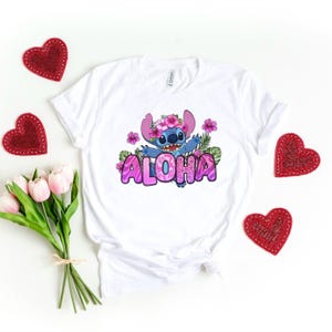 Puede incluir: Camiseta blanca con un personaje azul Stitch que lleva una corona de flores rosas y dice "Aloha" en letras rosas con un dise&ntilde;o floral tropical.