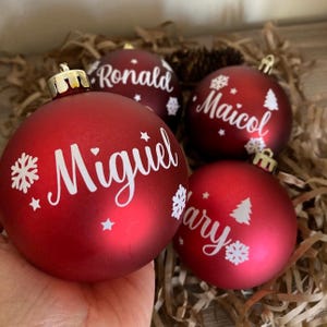 May include: Four red Christmas ornaments with white lettering and snowflake and star designs. The names "Miguel", "Ronald", "Maicol", and "Mary" are printed on the ornaments. Each ornament has a gold-colored cap.