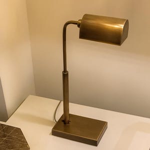 May include: A brass-toned desk lamp with a rectangular base and a cylindrical shade. The lamp has a slender, adjustable arm and a simple, modern design. The lamp is approximately 12 inches tall.