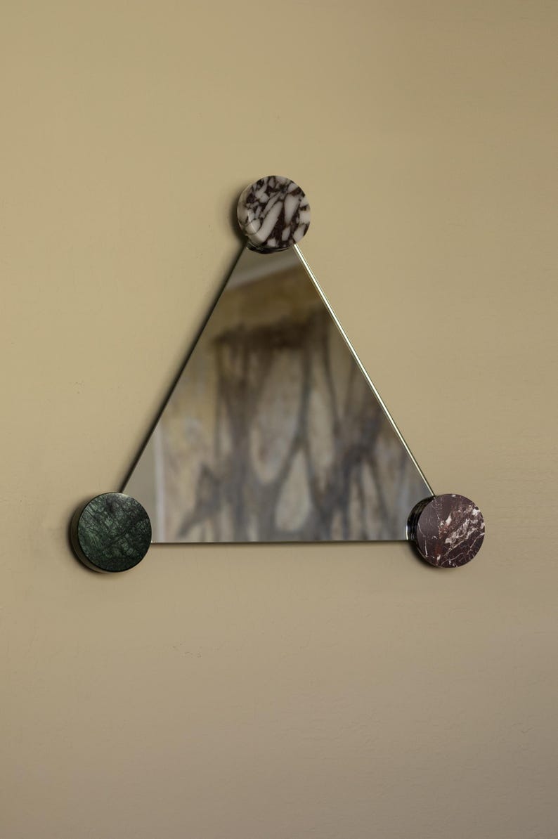 May include: A triangular mirror with a silver frame, held in place by three circular marble accents. The marble accents are white and black, green, and maroon. The mirror reflects a blurred image.