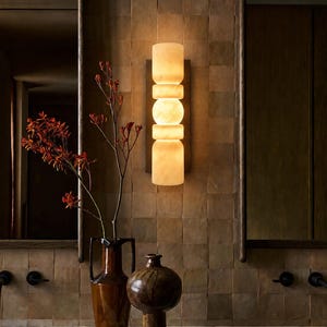 May include: A vertical wall sconce featuring stacked, cylindrical, and spherical translucent white stone segments that emit a warm, soft glow. The light fixture is mounted on a rectangular dark metal backplate against a tiled wall surface.