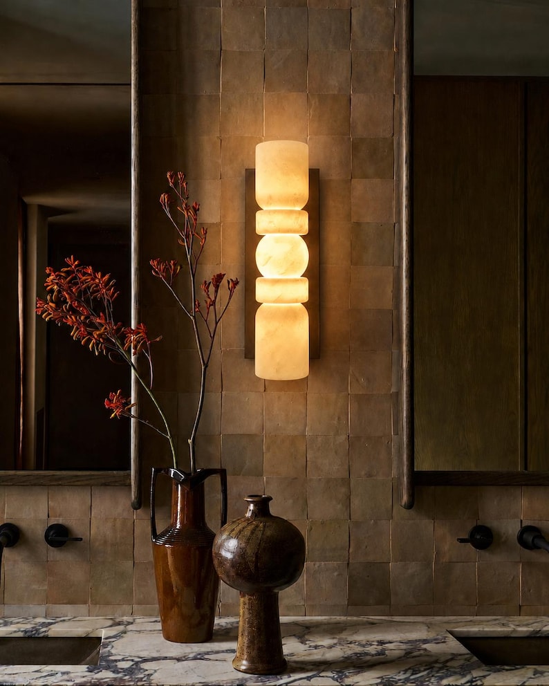 May include: A vertical wall sconce featuring stacked, cylindrical, and spherical translucent white stone segments that emit a warm, soft glow. The light fixture is mounted on a rectangular dark metal backplate against a tiled wall surface.