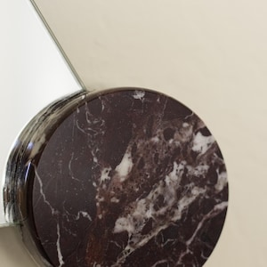 May include: A round, dark burgundy marble object with white veining, attached to a reflective, silver-toned surface. The marble has a polished finish. The background is a solid, light beige color.