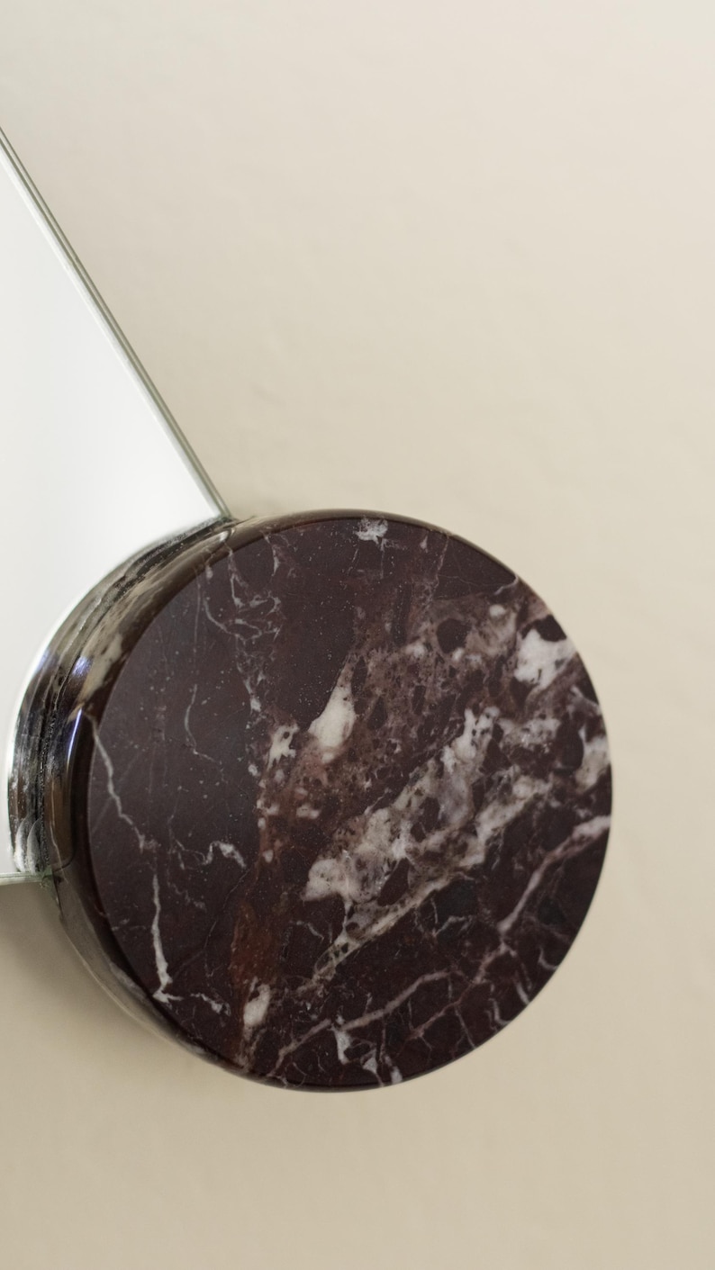 May include: A round, dark burgundy marble object with white veining, attached to a reflective, silver-toned surface. The marble has a polished finish. The background is a solid, light beige color.