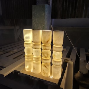 May include: Four cylindrical lamps made of translucent white stone with natural beige veining. Each lamp features a stacked design of three rounded segments and a smooth top section. The lamps emit a warm, soft glow from within, illuminating the stone.