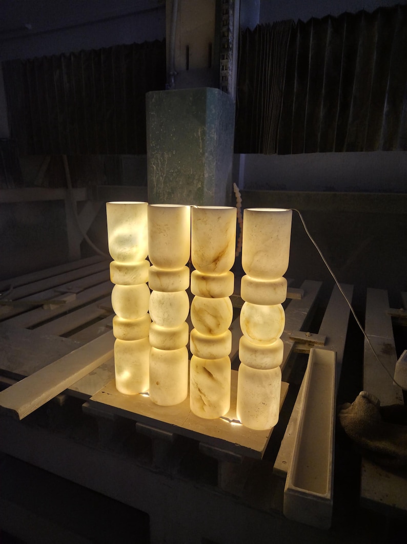 May include: Four cylindrical lamps made of translucent white stone with natural beige veining. Each lamp features a stacked design of three rounded segments and a smooth top section. The lamps emit a warm, soft glow from within, illuminating the stone.