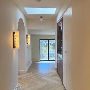 May include: A hallway featuring light-colored herringbone wood flooring, white walls, and a skylight. Two wall-mounted sconces with stacked cylindrical glass shades provide warm lighting. A dark-toned arched cabinet is built into the right wall.