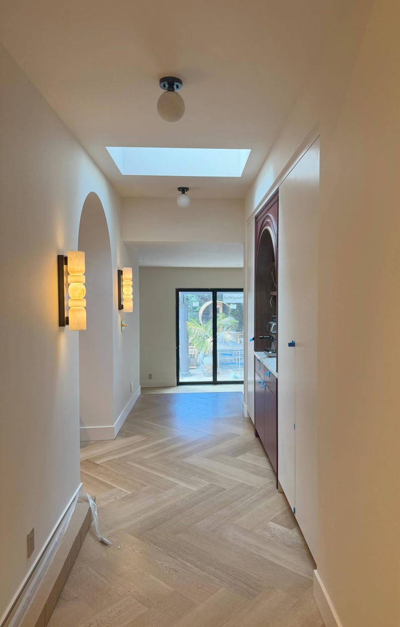 May include: A hallway featuring light-colored herringbone wood flooring, white walls, and a skylight. Two wall-mounted sconces with stacked cylindrical glass shades provide warm lighting. A dark-toned arched cabinet is built into the right wall.