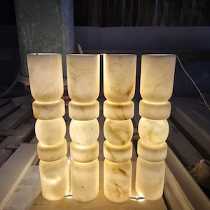 May include: Four cylindrical lamps made of translucent white stone with natural veining. Each lamp features a stacked design with three distinct rounded segments and a smooth finish, emitting a warm, soft glow from within the stone structure.