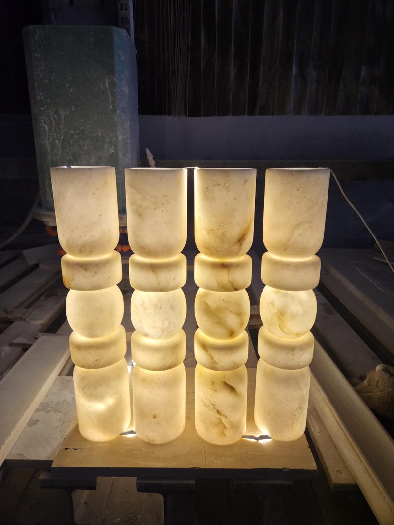 May include: Four cylindrical lamps made of translucent white stone with natural veining. Each lamp features a stacked design with three distinct rounded segments and a smooth finish, emitting a warm, soft glow from within the stone structure.