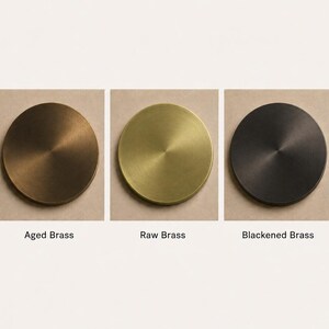 May include: Three circular metal discs displayed in a row, each featuring a different finish. The left disc is labeled Aged Brass, the center disc is labeled Raw Brass, and the right disc is labeled Blackened Brass. Each disc has a smooth surface.