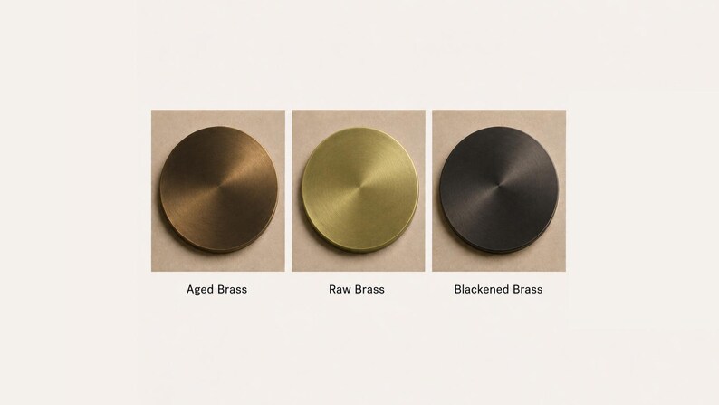 May include: Three circular metal discs displayed in a row, each featuring a different finish. The left disc is labeled Aged Brass, the center disc is labeled Raw Brass, and the right disc is labeled Blackened Brass. Each disc has a smooth surface.