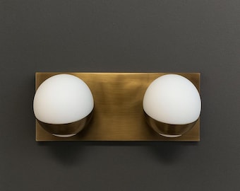May include: A wall-mounted light fixture featuring a rectangular brushed brass backplate and two spherical white glass globes. Each globe is partially encased in a matching brushed brass band that wraps around the lower portion of the glass.