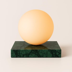 May include: A contemporary lamp with a large, spherical, peach-coloured light fixture on a dark green marble base. The base is a rectangular block with a veined pattern. The lamp is set against a neutral background.