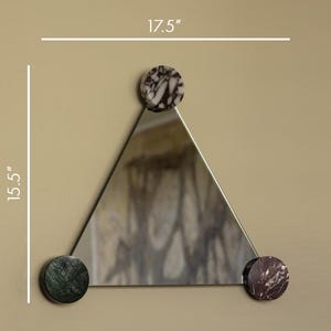 May include: A triangular mirror with a 17.5-inch width and 15.5-inch height. The mirror is accented with three circular marble pieces in shades of brown, green, and red. The mirror is mounted on a neutral-colored wall.