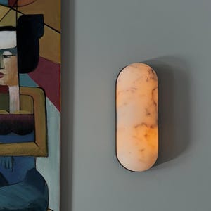 May include: A white marble oval wall sconce with a warm light emanating from inside. The sconce is mounted on a grey wall.