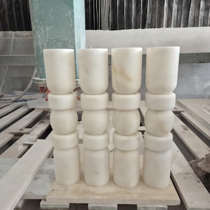 May include: Four vertical columns made of white marble, each composed of stacked cylindrical and rounded segments. The stone features subtle grey veining throughout the smooth, polished surface. These architectural elements are arranged in a single row.