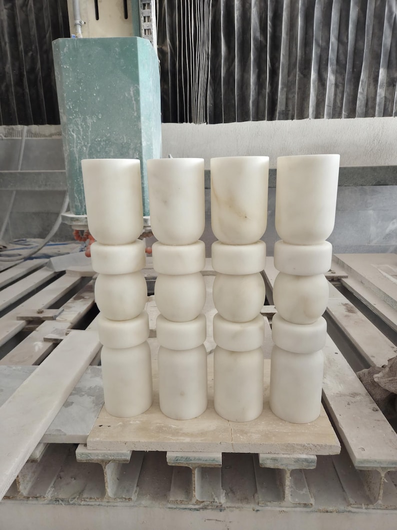 May include: Four vertical columns made of white marble, each composed of stacked cylindrical and rounded segments. The stone features subtle grey veining throughout the smooth, polished surface. These architectural elements are arranged in a single row.