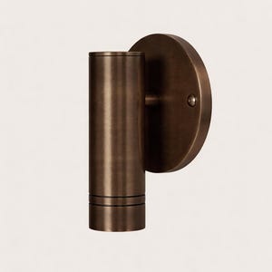 May include: A bronze-colored wall sconce with a cylindrical light fixture and a circular backplate. The sconce has a minimalist design, suitable for modern interiors. The fixture is approximately 6 inches tall.