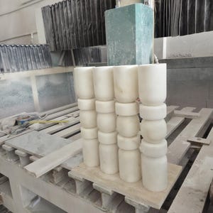May include: Four vertical white stone pillars arranged in a row, each featuring a series of stacked cylindrical and rounded segments. The pillars are crafted from a smooth, light-colored stone material with subtle natural veining throughout the surface.