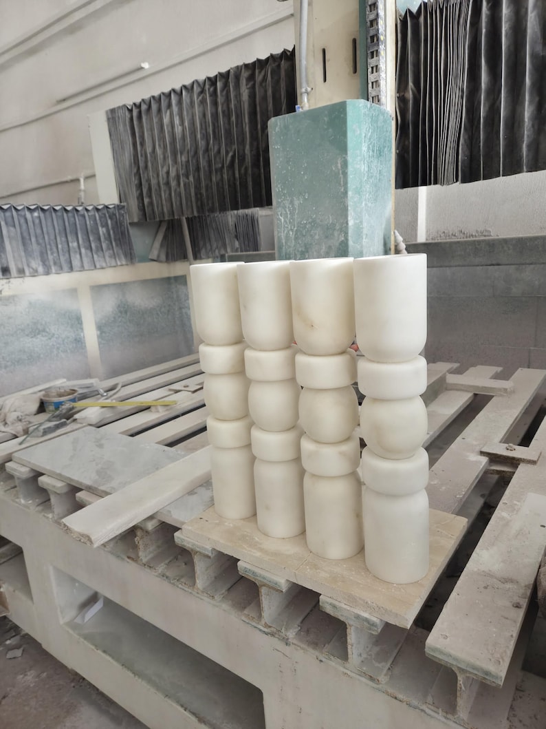 May include: Four vertical white stone pillars arranged in a row, each featuring a series of stacked cylindrical and rounded segments. The pillars are crafted from a smooth, light-colored stone material with subtle natural veining throughout the surface.