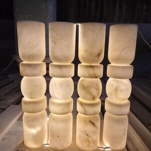 May include: Four cylindrical floor lamps made of translucent white stone with natural veining. Each lamp features a stacked design consisting of a tall base, a thick ring, a central sphere, another thick ring, and a tall top section, all illuminated.