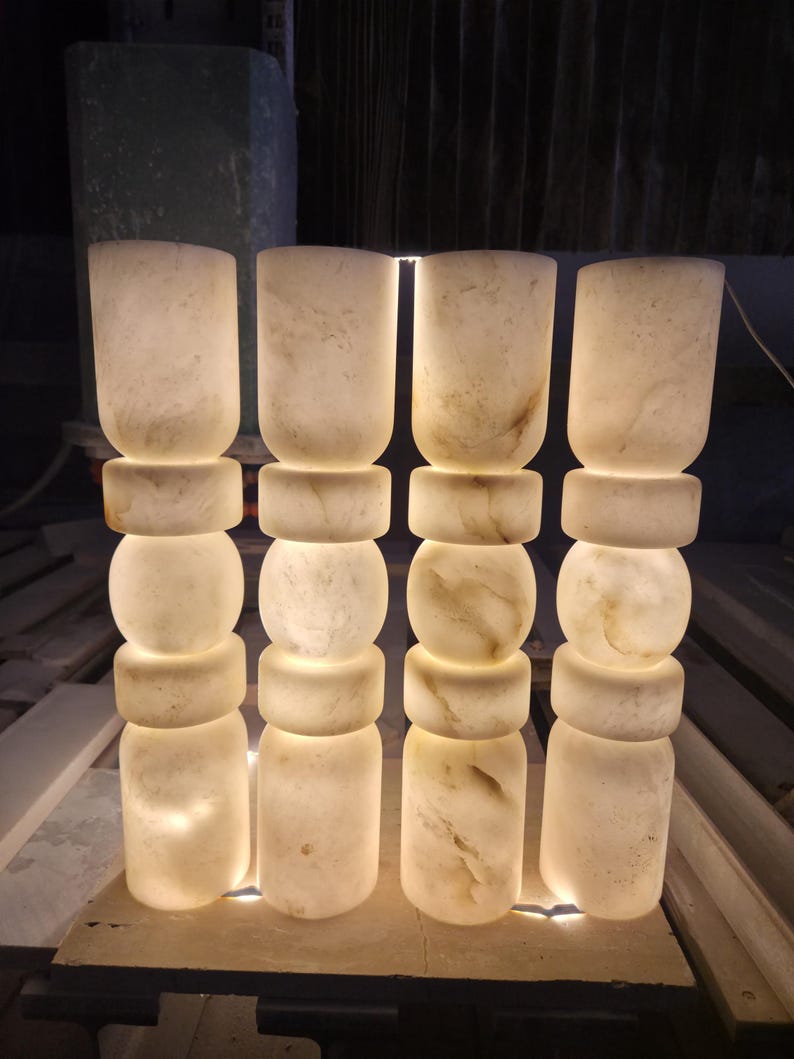 May include: Four cylindrical floor lamps made of translucent white stone with natural veining. Each lamp features a stacked design consisting of a tall base, a thick ring, a central sphere, another thick ring, and a tall top section, all illuminated.