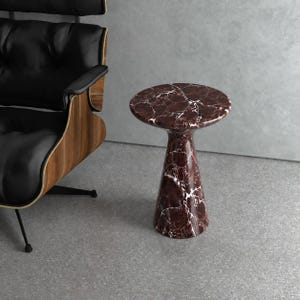 May include: A round side table crafted from deep red marble with prominent white veining. The table features a smooth, circular top supported by a tapered, conical base that widens towards the floor, showcasing a polished stone finish throughout.