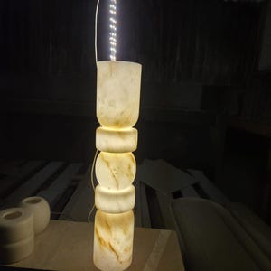 May include: A tall, cylindrical floor lamp crafted from translucent, cream-colored stone with natural brown veining. The lamp features a stacked, geometric design with alternating rounded and cylindrical segments, illuminated from within by an LED strip.
