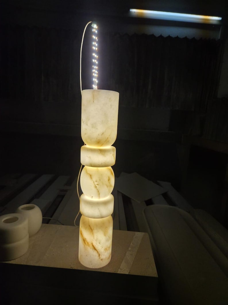 May include: A tall, cylindrical floor lamp crafted from translucent, cream-colored stone with natural brown veining. The lamp features a stacked, geometric design with alternating rounded and cylindrical segments, illuminated from within by an LED strip.