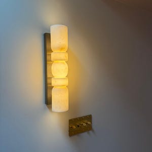 May include: A vertical wall sconce featuring a stack of five cylindrical and spherical off-white stone segments that emit a warm, soft glow. The light fixture is mounted on a dark rectangular base, positioned above a metallic gold light switch plate.