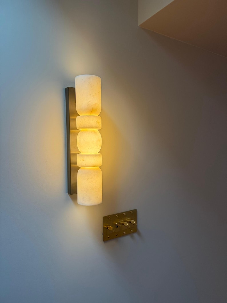 May include: A vertical wall sconce featuring a stack of five cylindrical and spherical off-white stone segments that emit a warm, soft glow. The light fixture is mounted on a dark rectangular base, positioned above a metallic gold light switch plate.