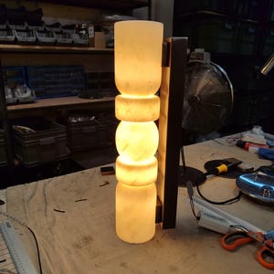 May include: A vertical wall sconce featuring a stack of five cylindrical and spherical translucent white stone segments that emit a warm, soft glow. The light fixture is mounted to a dark rectangular metal backplate with visible wiring.