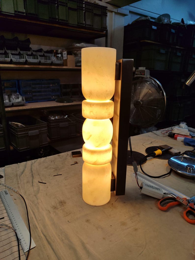 May include: A vertical wall sconce featuring a stack of five cylindrical and spherical translucent white stone segments that emit a warm, soft glow. The light fixture is mounted to a dark rectangular metal backplate with visible wiring.