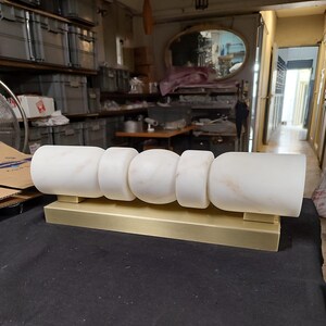 May include: A horizontal wall light fixture featuring a long, rectangular brushed gold metal base supporting five white marble segments. The marble pieces include two large cylinders at the ends, two smaller rings, and a central sphere, all aligned.