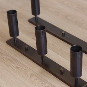 May include: Two dark brown metal candle holders, each featuring a long rectangular base with three vertical cylindrical sockets attached by short stems and small circular accents positioned between the sockets on the flat base surface.