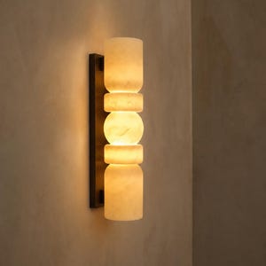 May include: A wall sconce featuring a vertical arrangement of five stacked, translucent, off-white stone segments with a marbled texture. The segments are mounted on a thin, rectangular, dark brown metal backplate, emitting a warm, soft glow.