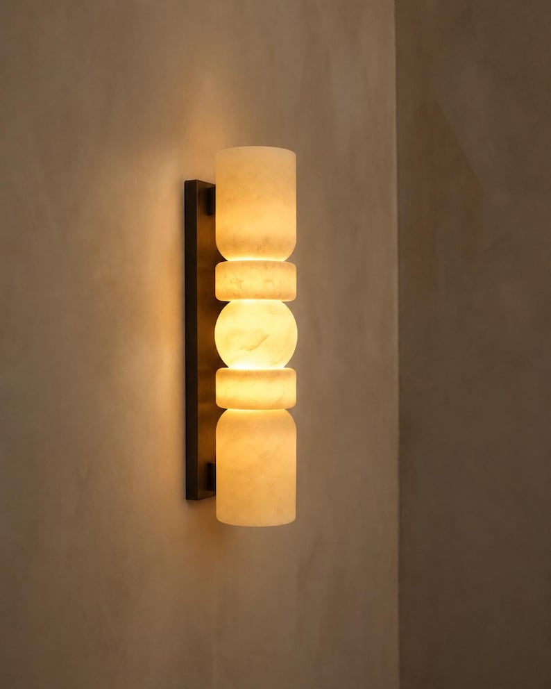 May include: A wall sconce featuring a vertical arrangement of five stacked, translucent, off-white stone segments with a marbled texture. The segments are mounted on a thin, rectangular, dark brown metal backplate, emitting a warm, soft glow.