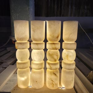 May include: Four cylindrical floor lamps made of translucent white stone with natural beige veining. Each lamp features a stacked design with alternating smooth cylindrical and rounded sections, emitting a warm, soft glow from within the stone.