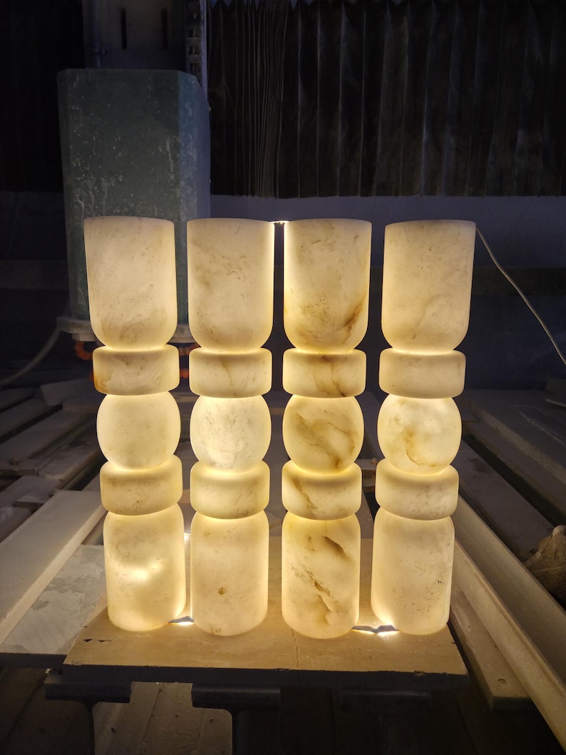 May include: Four cylindrical floor lamps made of translucent white stone with natural beige veining. Each lamp features a stacked design with alternating smooth cylindrical and rounded sections, emitting a warm, soft glow from within the stone.